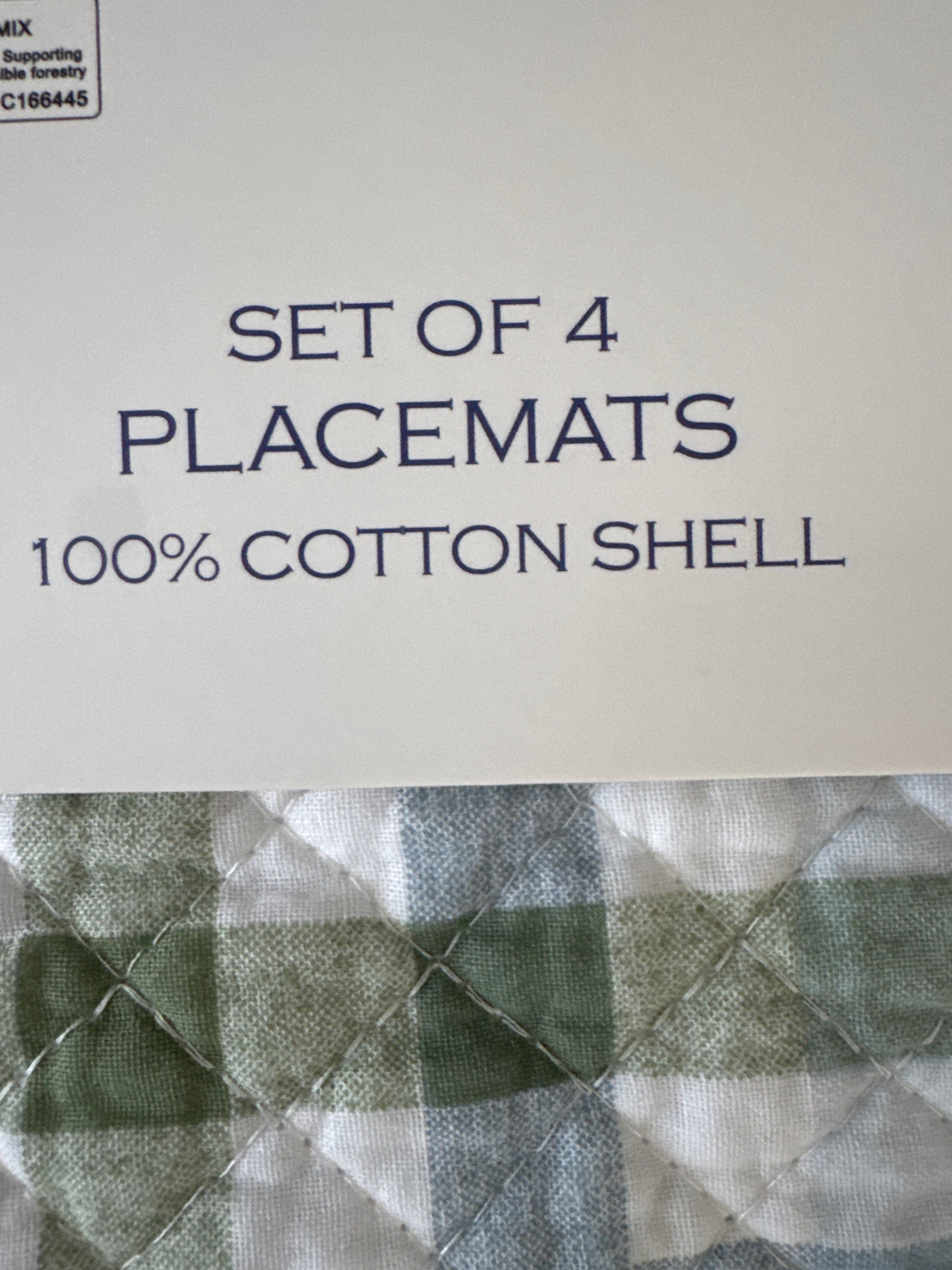 Close-up of the label on a set of 4 cotton placemats, displaying the text "SET OF 4 PLACEMATS 100% COTTON SHELL." The background shows a green and white plaid pattern on the placemats
